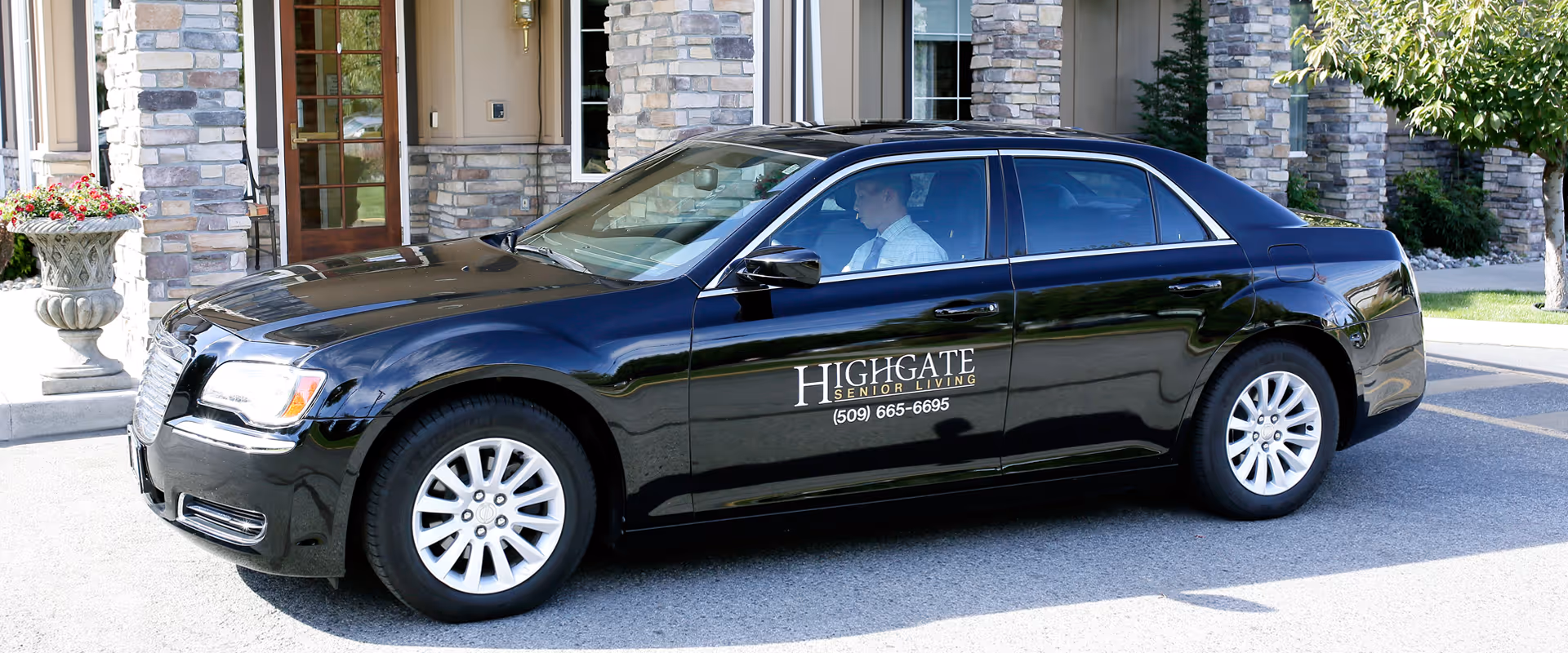 A black sedan car parked in front of a building entrance with stone pillars and a wooden door. The car has the logo and contact number of Highgate Senior Living on its side. A person is seated inside the car.