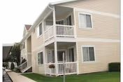 Two-story beige apartment building with covered first- and second-floor porches, white trim, and a small front lawn.