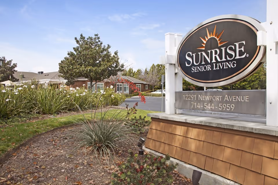 Outdoor view of the entrance sign for Sunrise Senior Living at 12291 Newport Avenue, surrounded by landscaping with trees, bushes, and flowers, with a building visible in the background under a clear sky.