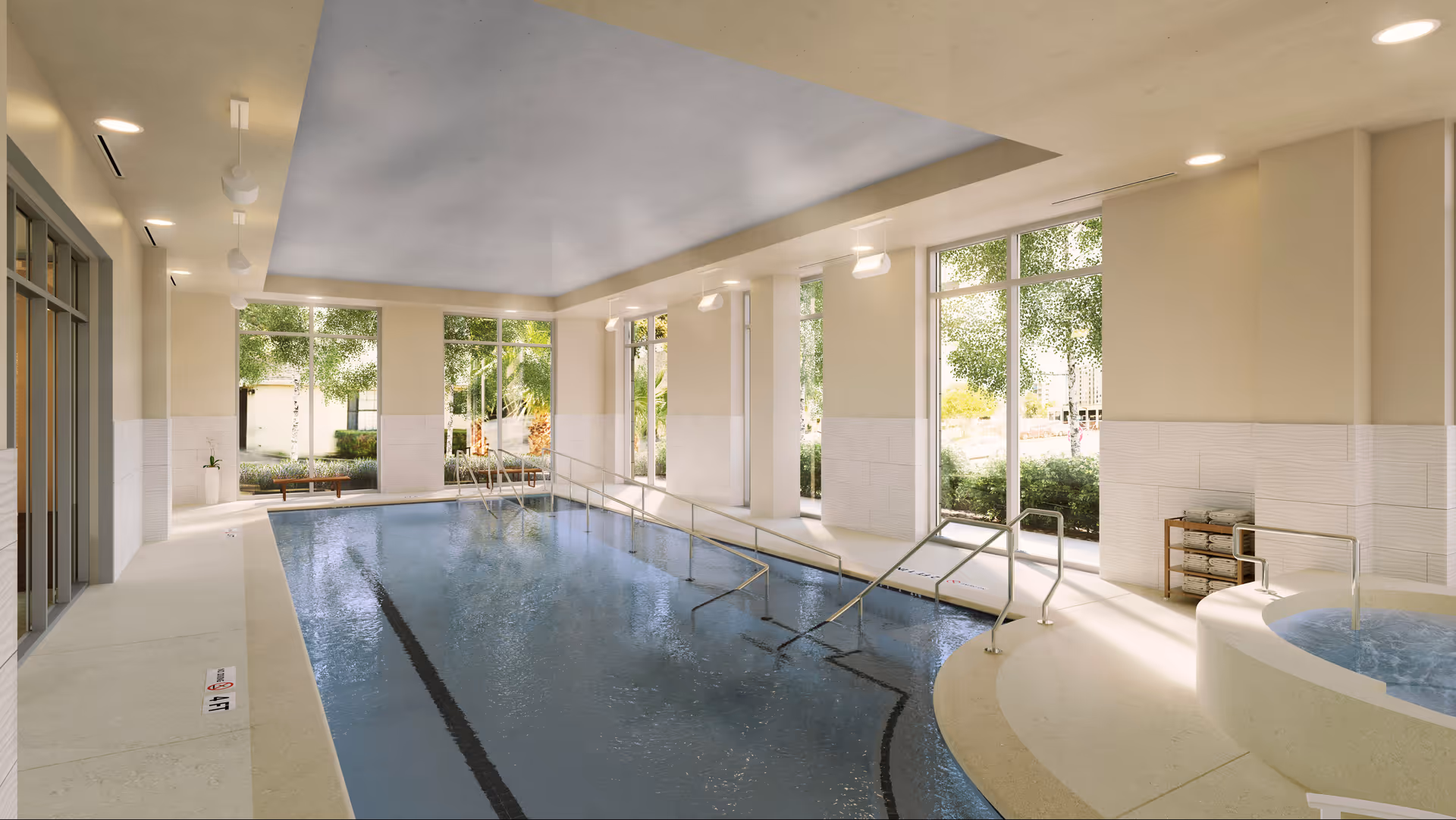 Indoor swimming pool area with large windows letting in natural light, handrails for accessibility, and a small circular hot tub on the right side. Outside greenery is visible through the windows.