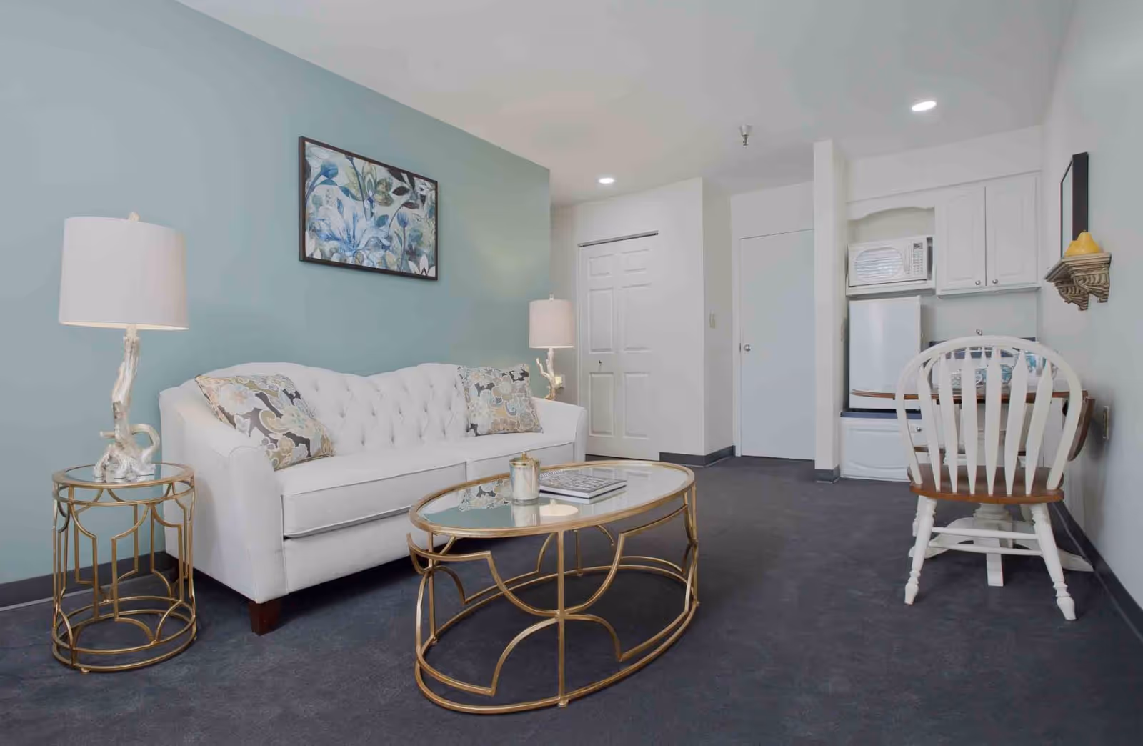A cozy living area with a white tufted sofa adorned with patterned pillows against a light blue wall. A glass-top coffee table with a gold metal frame sits in front of the sofa, accompanied by a matching side table with a lamp. In the background, there is a small kitchenette with a microwave, mini fridge, and a white wooden chair at a small table. The room has gray carpeting and white walls.