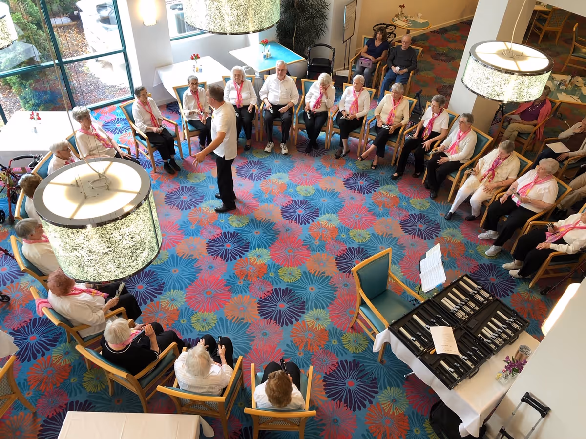 A group of elderly people seated in a large circle in a brightly lit room with colorful floral carpet. A man stands in the middle, seemingly leading an activity. The room has large windows, modern hanging lamps, and tables with chairs around the perimeter.