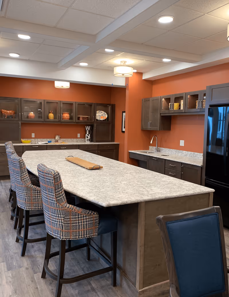 A modern kitchen area with a large marble countertop island surrounded by four plaid upholstered chairs. The kitchen features dark wood cabinets, a sink, a black refrigerator, and decorative items on the shelves against a burnt orange wall. The ceiling has recessed lighting and two hanging light fixtures.