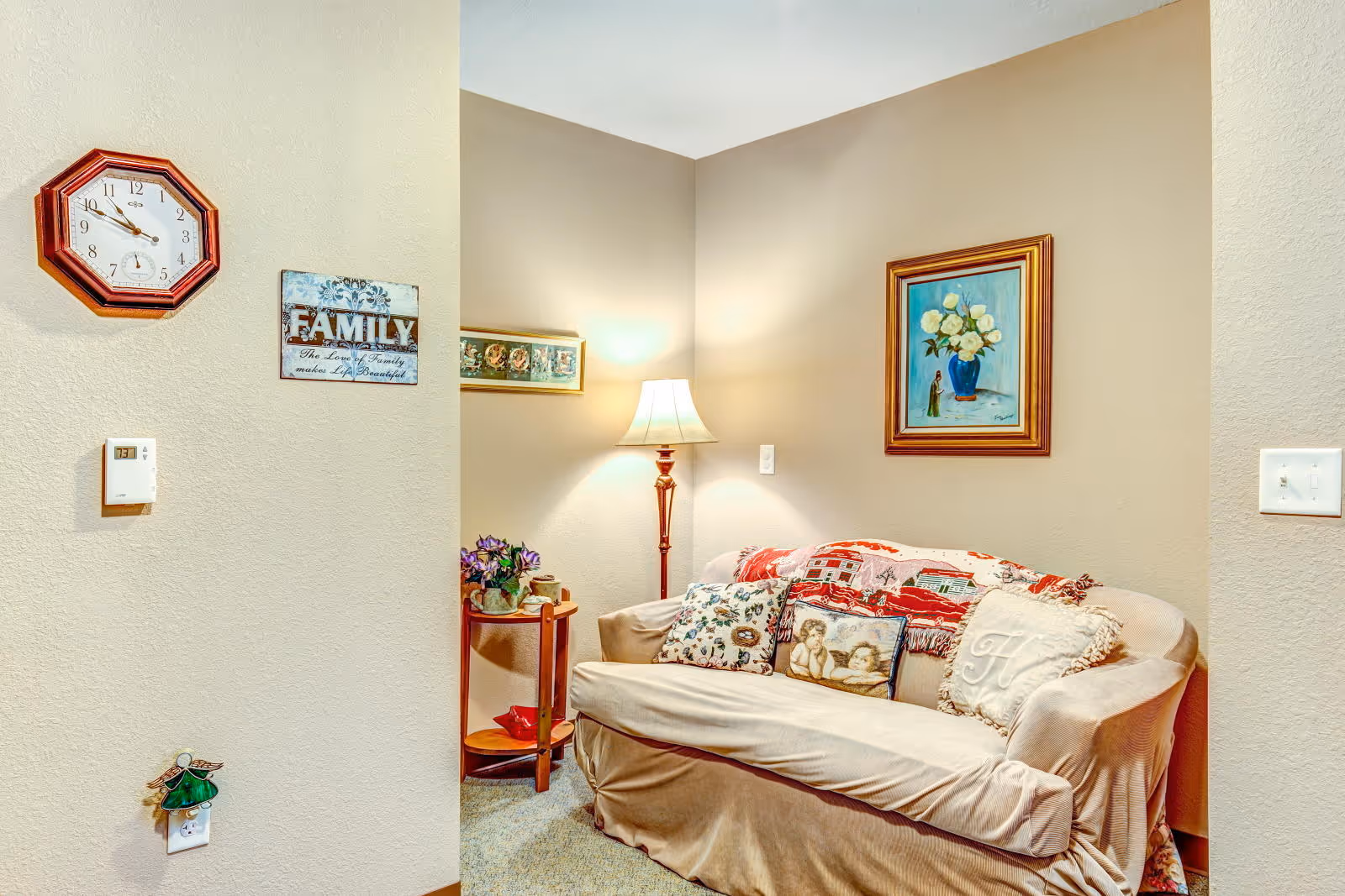 A cozy corner of a living room with a beige couch adorned with decorative pillows and a throw blanket. A floor lamp stands behind the couch, illuminating the space. On the wall above the couch is a framed painting of white flowers in a blue vase. To the left, a small round wooden side table holds a potted plant and other small items. A wall clock and a decorative sign that reads 'FAMILY The love of a family makes life beautiful' are mounted on the adjacent wall.