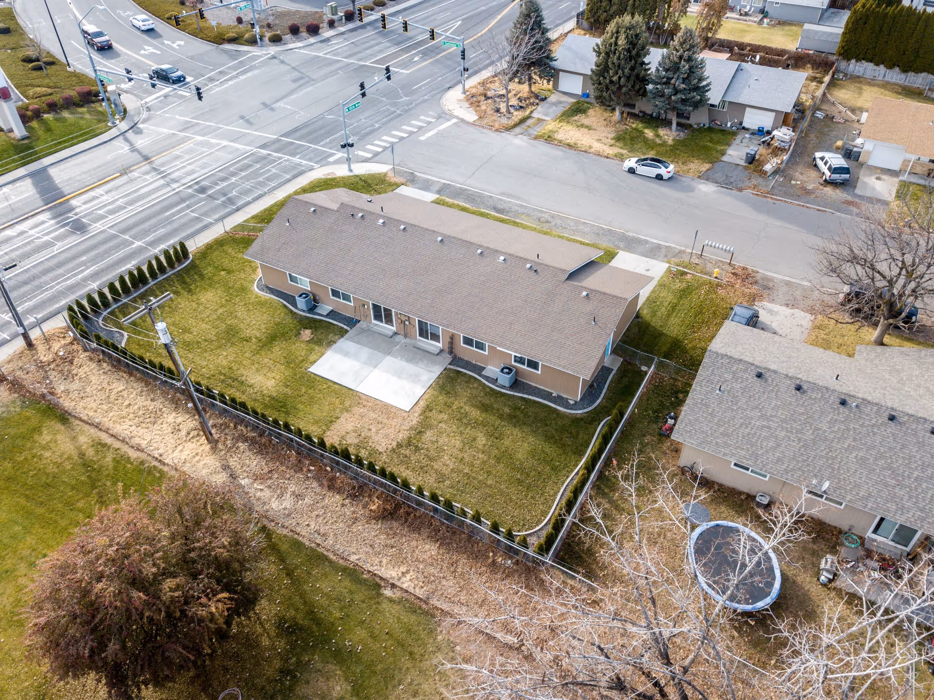 Aerial view of a single-story residential building with a fenced yard, located at the corner of two streets with traffic lights. The building has a brown roof and beige exterior walls, with a concrete patio area in the backyard. Surrounding the property are neighboring houses, trees, and a grassy area.