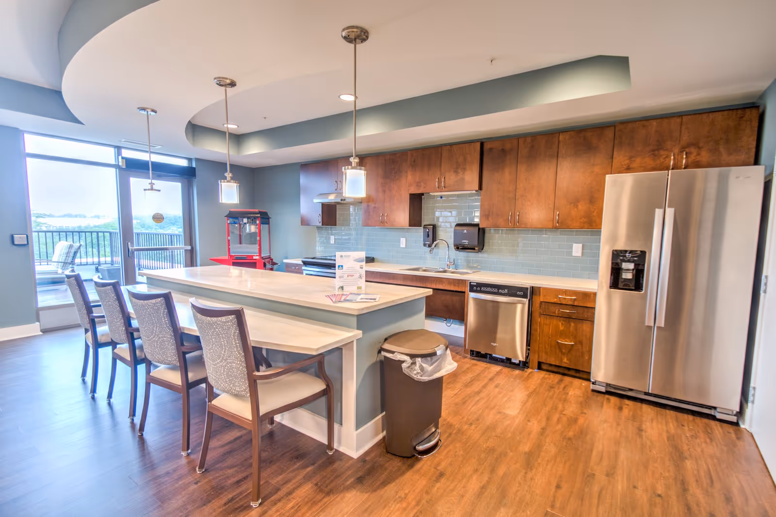 A modern kitchen area in a senior living facility featuring wooden cabinets, stainless steel refrigerator and dishwasher, a large island with seating for four, pendant lighting, and a popcorn machine near a glass door leading to an outdoor balcony.