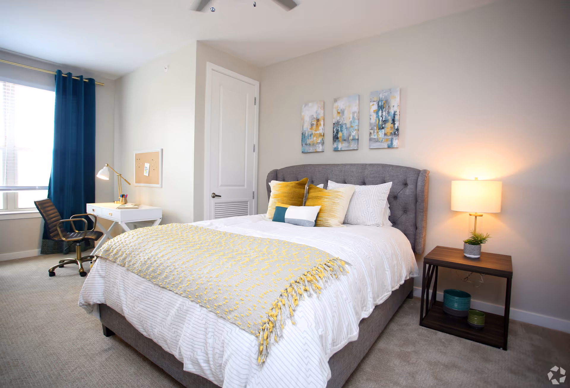 A bright and neatly arranged bedroom featuring a gray upholstered bed with white and yellow pillows and a yellow throw blanket. There is a wooden nightstand with a lamp and small plant on the right side of the bed. On the left side, there is a white desk with a gold lamp and a black office chair near a window with dark blue curtains. Three abstract paintings hang on the wall above the bed.