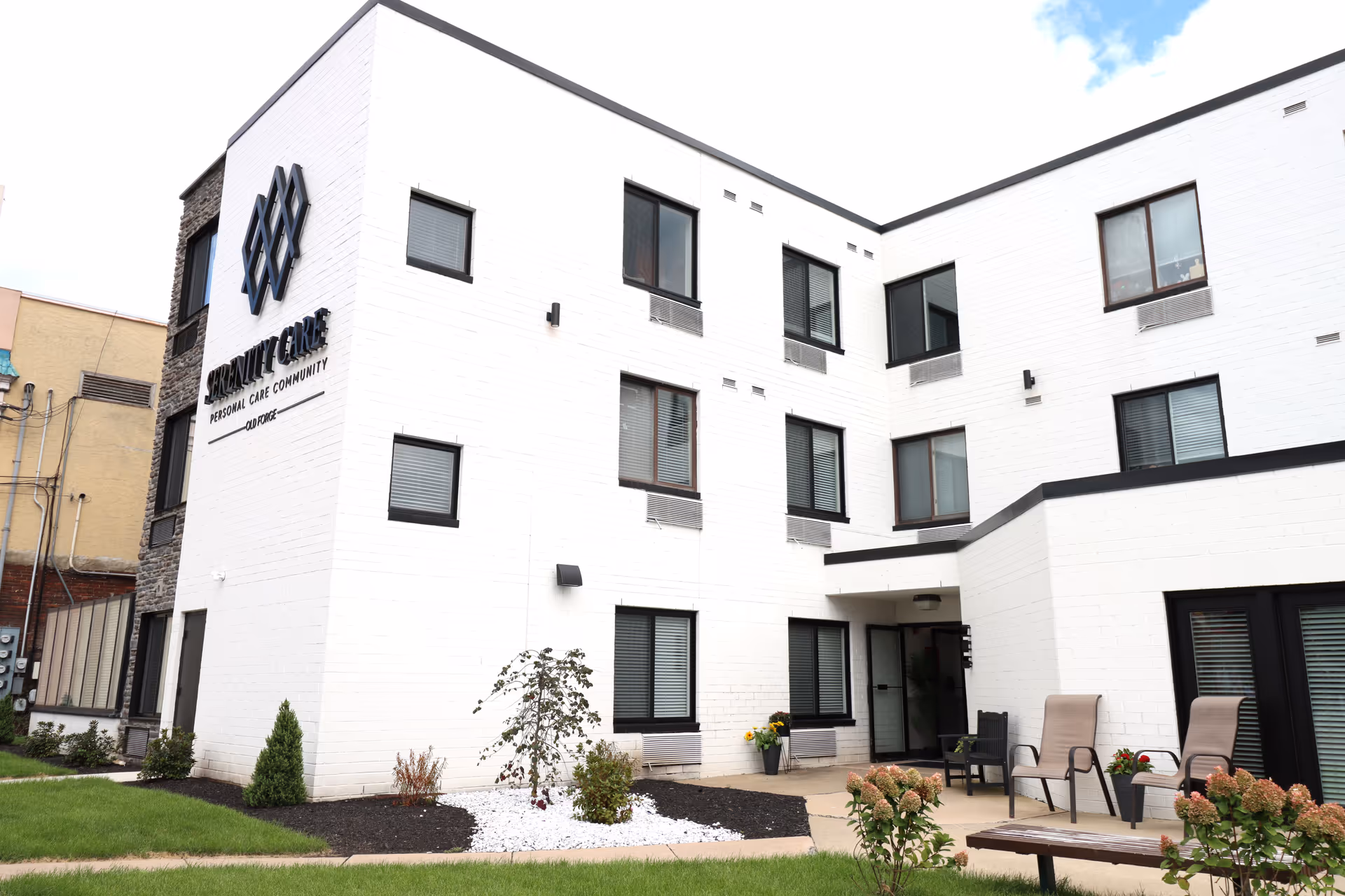 Exterior view of a white three-story building with multiple windows and a small landscaped garden area with chairs and plants in front. The building has a sign that reads 'Serenity Care Personal Care Community Old Forge'.