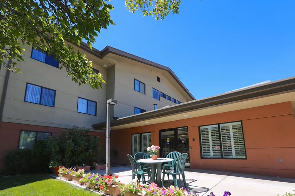 Outdoor patio area at St. Joseph Senior Living with a round table and four green chairs. The table has a potted plant with pink flowers. The patio is surrounded by flower beds with colorful flowers and greenery. The building exterior is visible with multiple windows and a clear blue sky above.