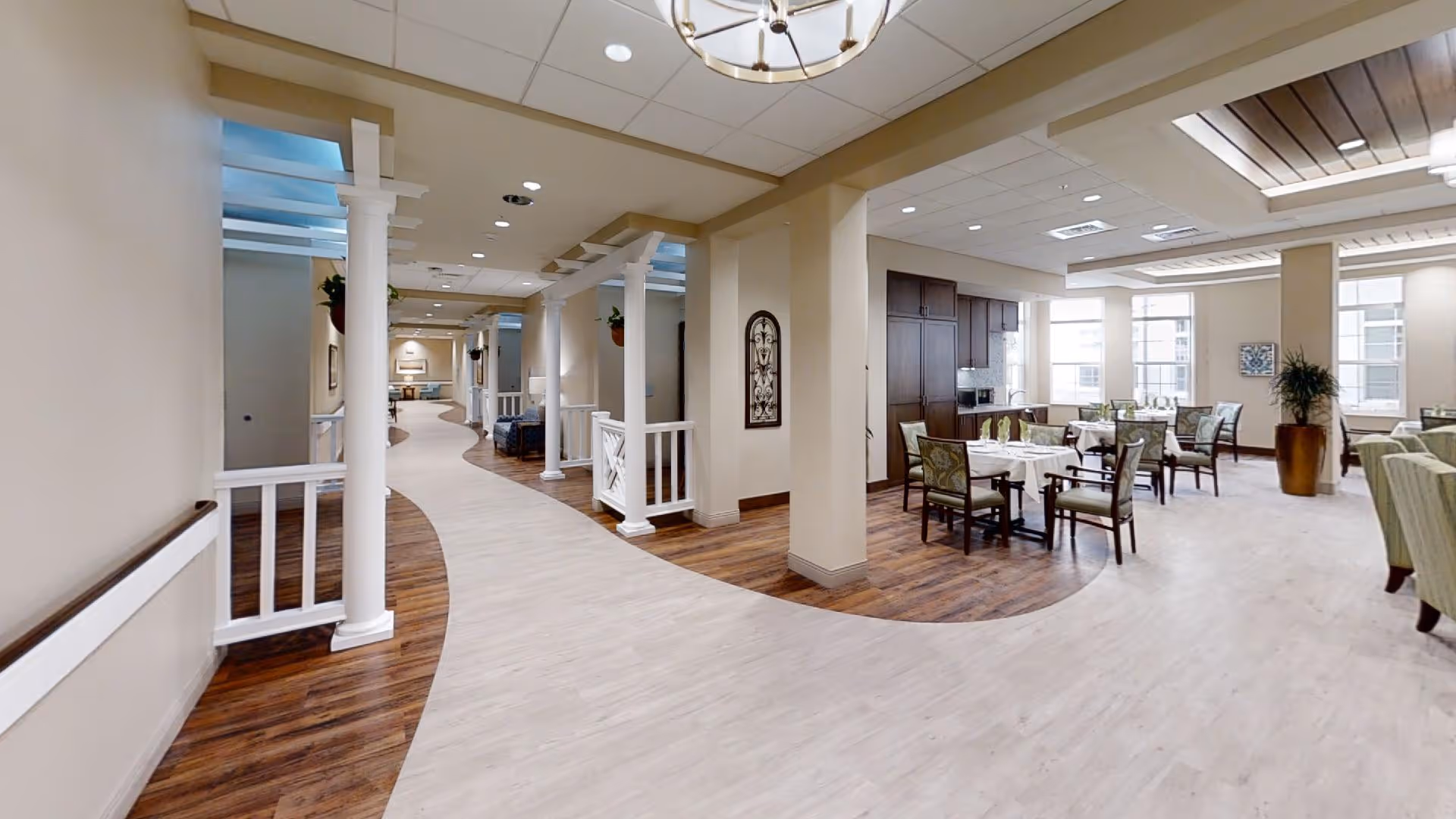 Interior view of a senior living facility showing a wide hallway with white columns and a curved light-colored floor. To the right, there is a dining area with tables and chairs set for a meal, large windows letting in natural light, and wood flooring under the dining space. The ceiling has recessed lighting and decorative fixtures.