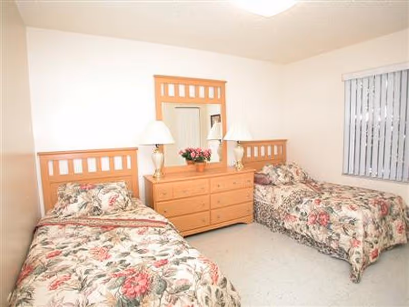 A bedroom with two twin beds featuring floral bedspreads and matching pillows. Between the beds is a wooden dresser with six drawers, topped with two table lamps and a flower pot with pink flowers. A large mirror is mounted on the wall above the dresser. A window with vertical blinds is on the right side of the room.