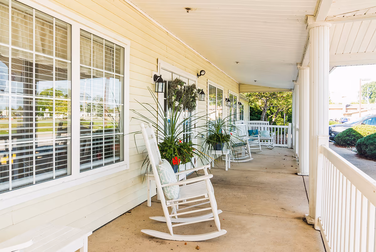 Covered porch area with white rocking chairs and potted plants along the wall of a building with large windows and cream-colored siding. The porch overlooks a parking area with bushes and trees in the background.