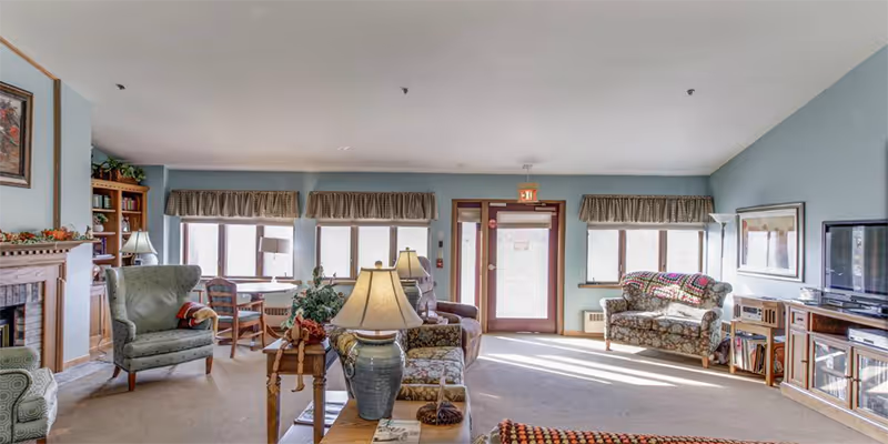 Spacious senior living lounge with sofas, chairs, lamps, a TV, and large windows.