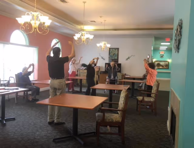 A group of elderly people and an instructor participate in a seated exercise or stretching activity in a well-lit room with large windows, chandeliers, and tables with chairs arranged around the space.