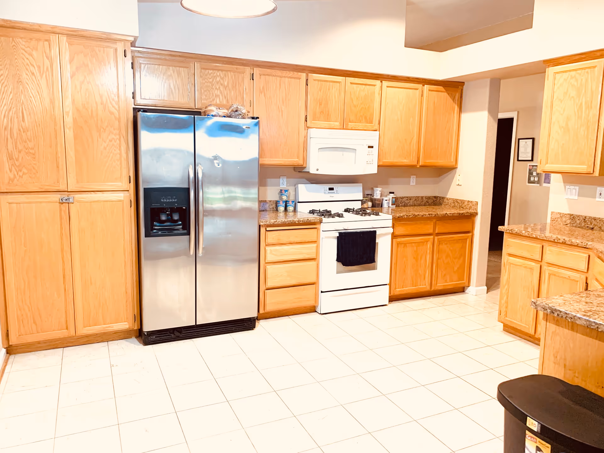A bright kitchen with wooden cabinets, a stainless steel refrigerator, a white gas stove with a microwave above it, granite countertops, and a tiled floor. There is a black trash bin in the corner and some canned goods on the counter.
