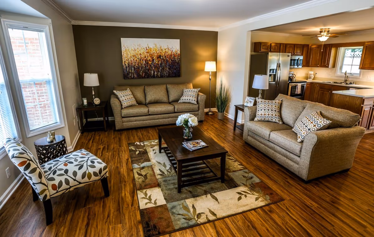 A cozy living room with two beige sofas adorned with patterned cushions, a wooden coffee table with a flower vase and magazines, a patterned armchair, two side tables with lamps, a floor lamp, a large window with blinds, and a kitchen area with wooden cabinets and stainless steel appliances in the background.