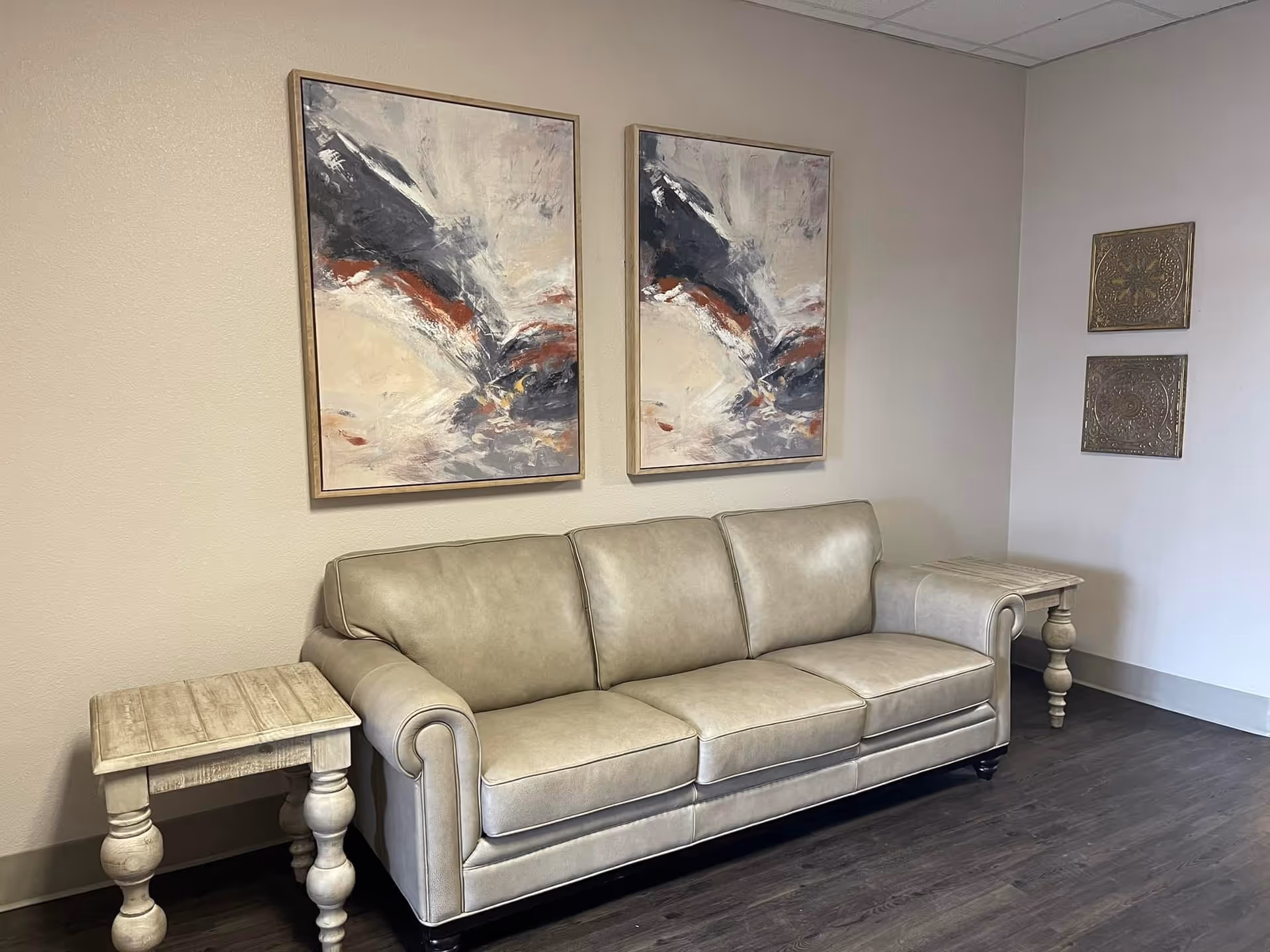 A beige leather sofa with rolled arms is positioned against a light-colored wall. On either side of the sofa are matching wooden side tables with turned legs. Above the sofa hang two abstract paintings with shades of gray, white, and rust. To the right on the adjacent wall are two decorative square metal wall hangings. The floor is dark wood.