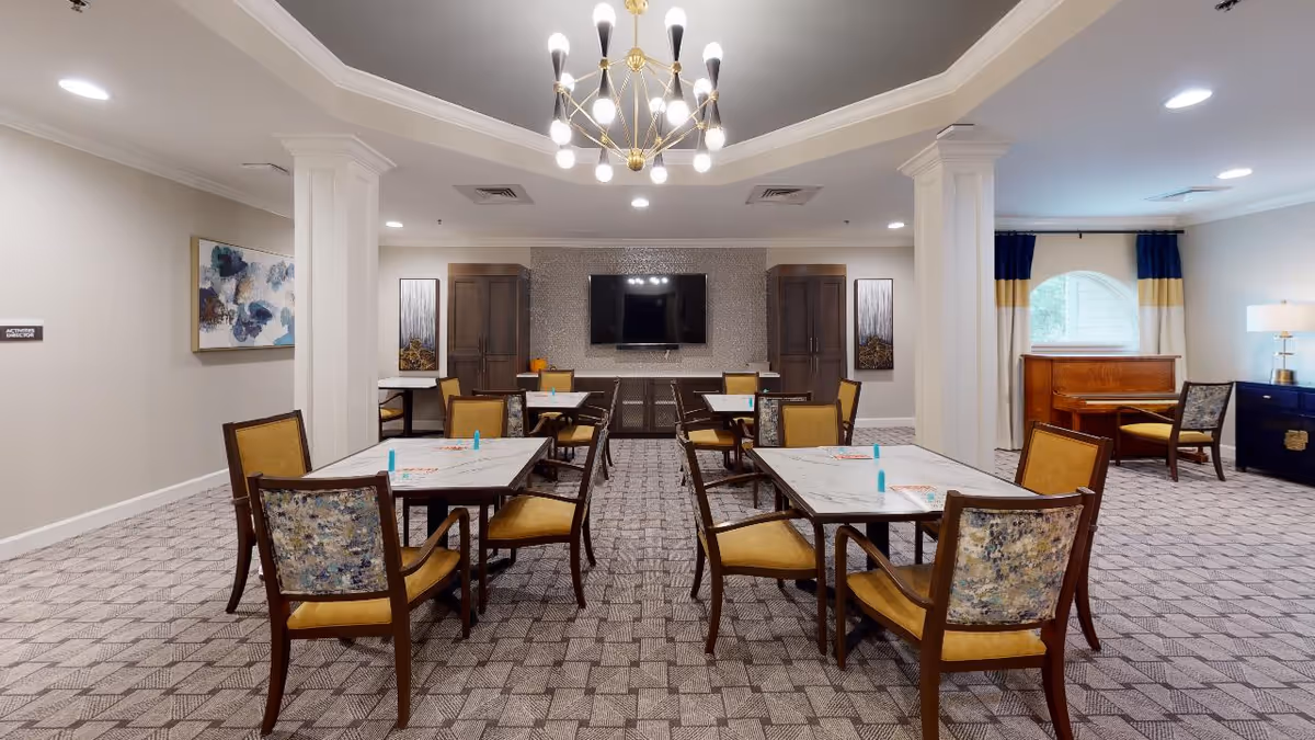 Communal dining/activity room with several tables and chairs, a chandelier, a wall-mounted TV, and a piano.