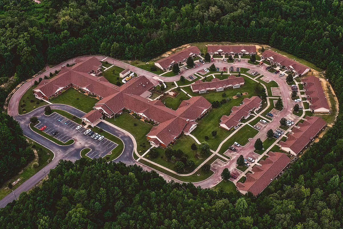 Aerial view of a senior living campus with multiple connected red-roof buildings, parking lots, walkways, and surrounding trees.