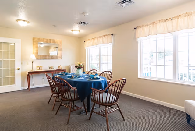 A bright dining room with a rectangular table covered with a blue tablecloth and set with plates and napkins. Six wooden chairs surround the table. The room has beige walls, two large windows with light curtains, a wooden side table with a lamp and decorative items, and a large mirror hanging above the side table.