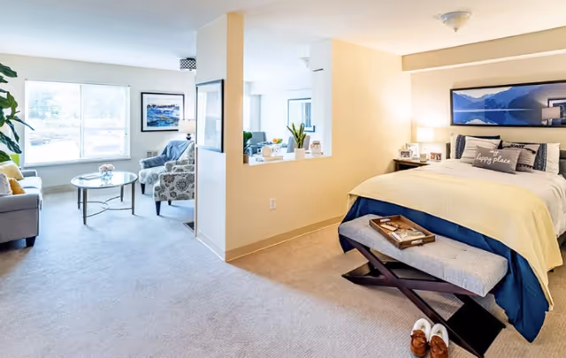 Bright studio-style senior living apartment with a bed on the right and a seating area with chairs and a coffee table by a large window on the left.