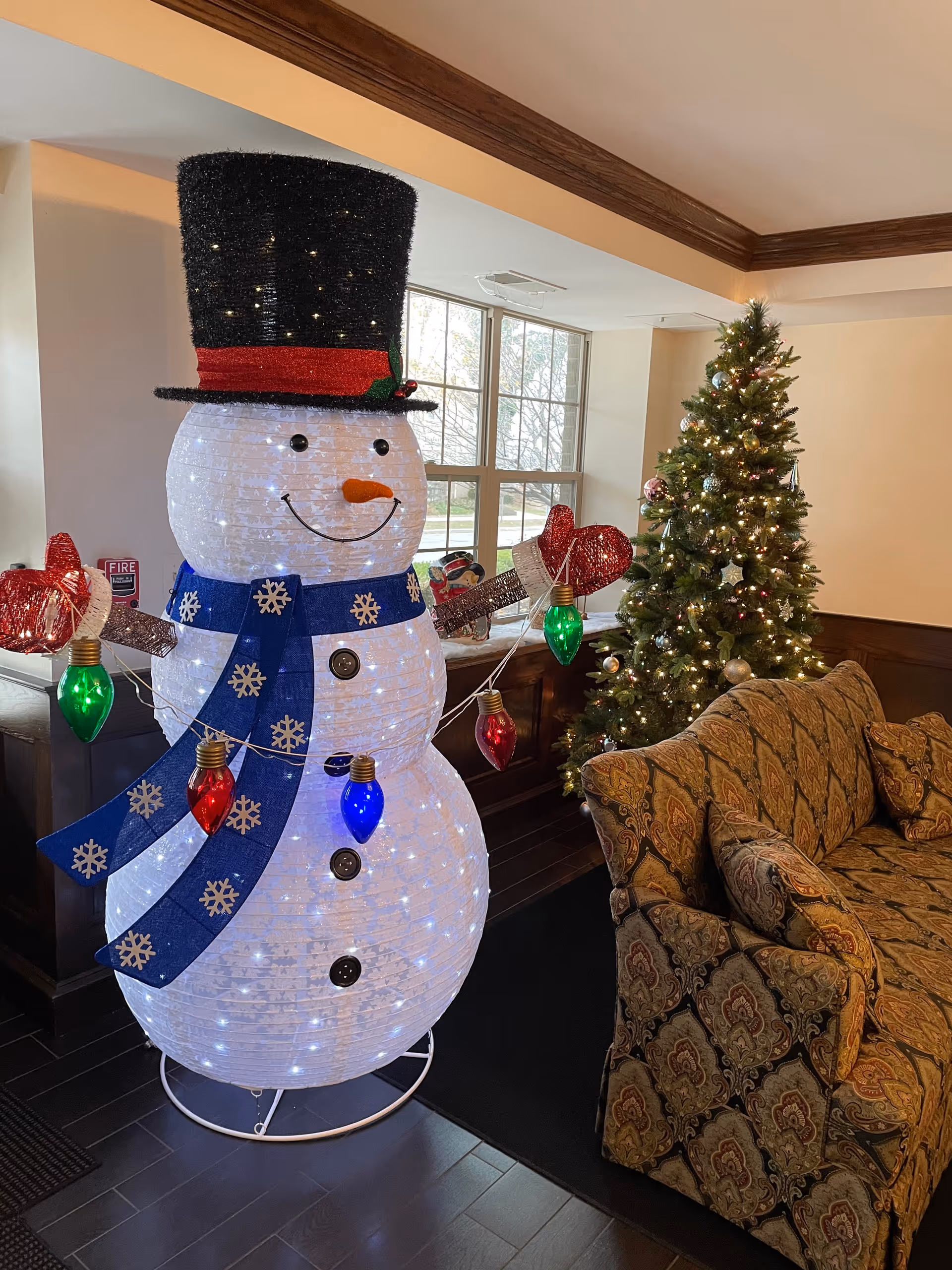 A festive indoor scene featuring a large illuminated snowman decoration wearing a black top hat with a red band and a blue scarf with snowflakes. The snowman is adorned with colorful Christmas light bulbs. Behind it is a decorated Christmas tree with lights and ornaments. To the right, there is a patterned sofa with cushions. Large windows in the background let in natural light.