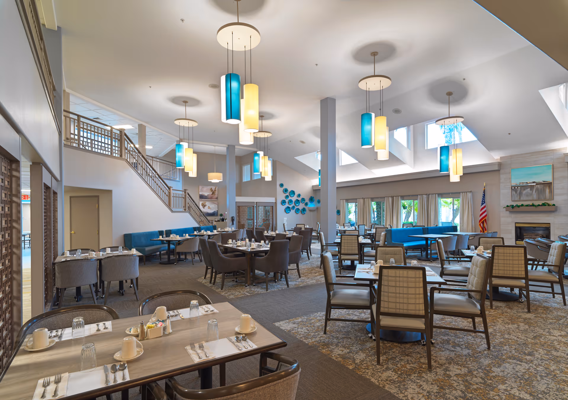Spacious, well-lit communal dining room with multiple set tables and chairs, pendant lights, a fireplace, and a staircase.