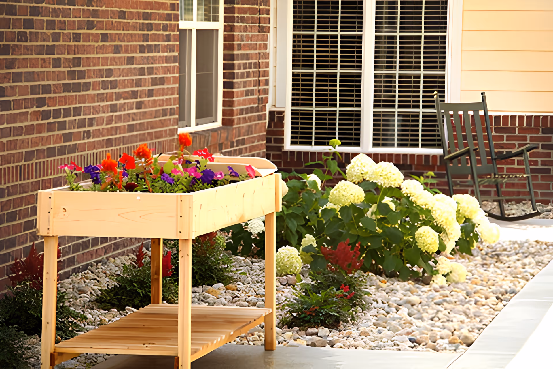 Outdoor patio area with a wooden raised garden bed filled with colorful flowers, a brick wall with windows, white flowering bushes, and a green rocking chair on a stone-covered ground.