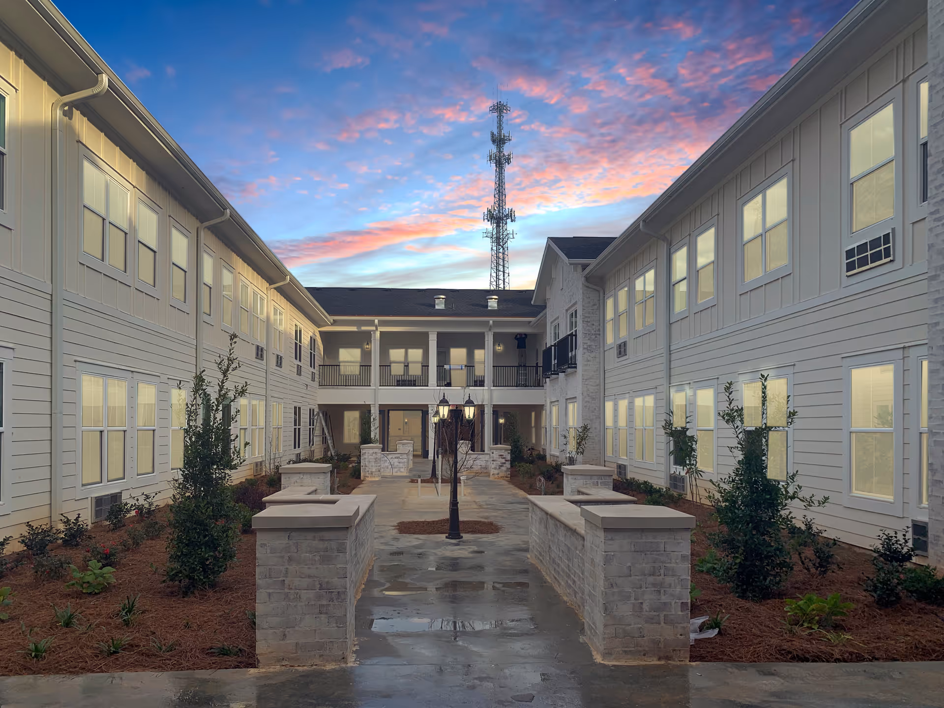 Outdoor courtyard area of The Blake at Biloxi facility during sunset, featuring a paved walkway lined with low brick walls and small landscaped plants, flanked by two-story buildings with multiple windows and a tall communication tower in the background under a colorful sky.