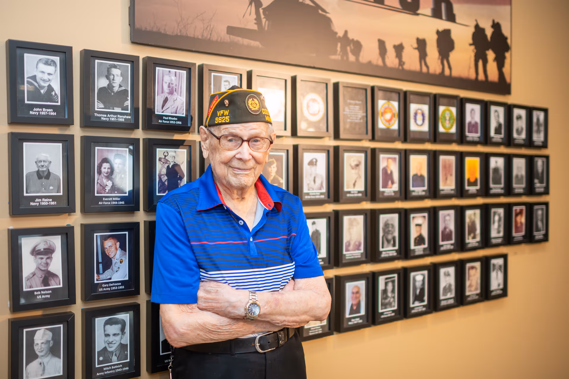 An elderly man wearing a blue striped polo shirt and a VFW military cap stands with his arms crossed in front of a wall displaying framed photographs and plaques honoring veterans.