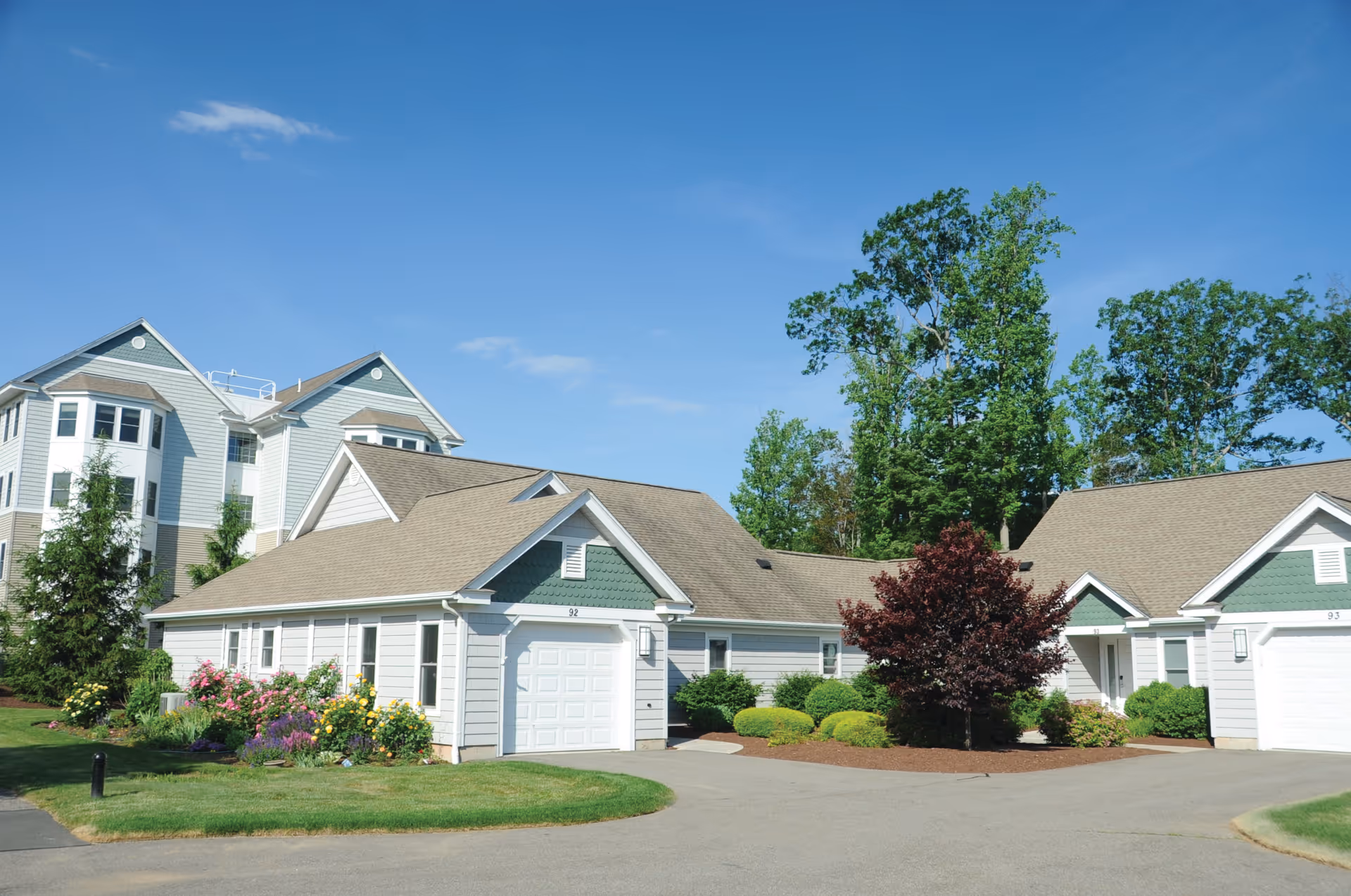 Exterior view of a senior living facility with light gray buildings featuring green accents, surrounded by well-maintained landscaping including colorful flowers, bushes, and trees under a clear blue sky.