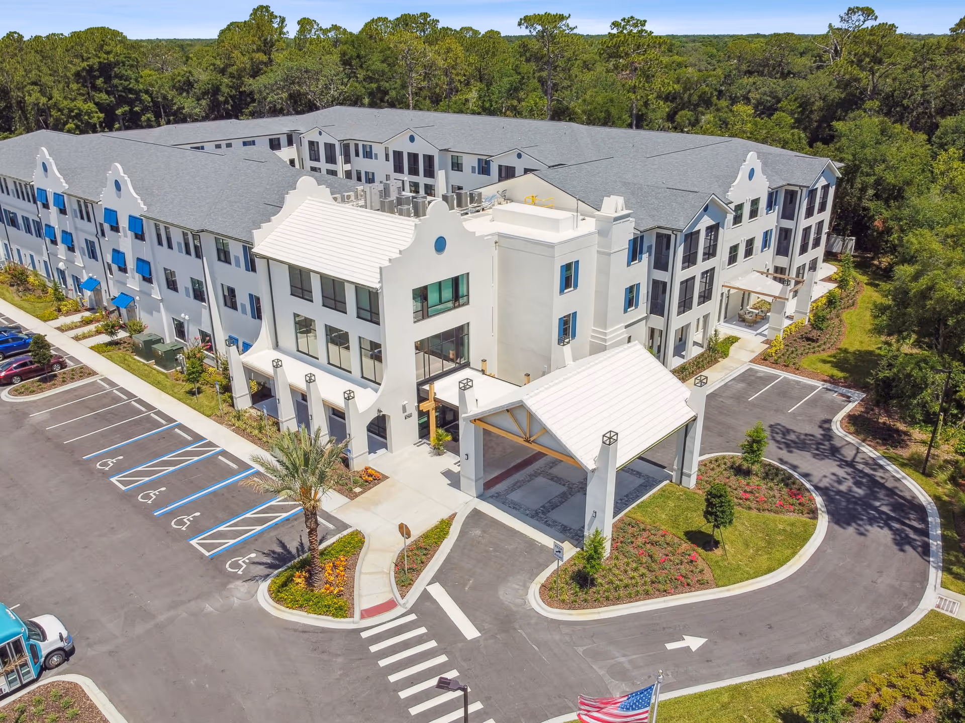 Aerial view of a three-story white senior living building with a covered entrance, parking spaces, and surrounding trees.