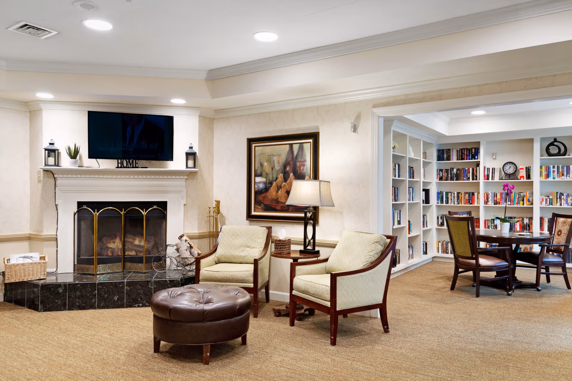 A cozy living room area with two cushioned armchairs and a round leather ottoman in front of a white fireplace with a black marble hearth. Above the fireplace is a mounted flat-screen TV and decorative items including lanterns and a small plant. To the right, there is a side table with a lamp and a framed painting on the wall. In the background, an adjoining room features built-in white bookshelves filled with books and a round table with chairs, creating a library or reading nook atmosphere.