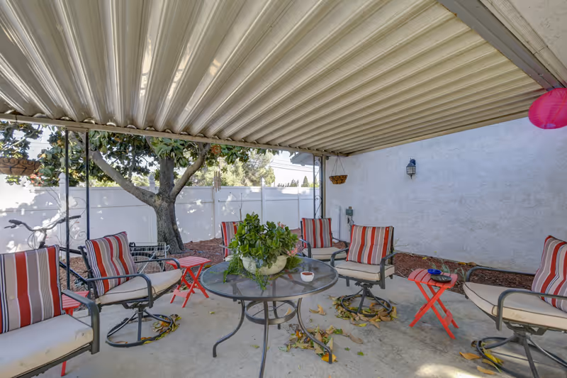 Covered outdoor patio area with a round glass table surrounded by six cushioned chairs with red and white striped pillows. There are two small red side tables, a potted plant on the table, and a tree visible outside the white fence in the background.