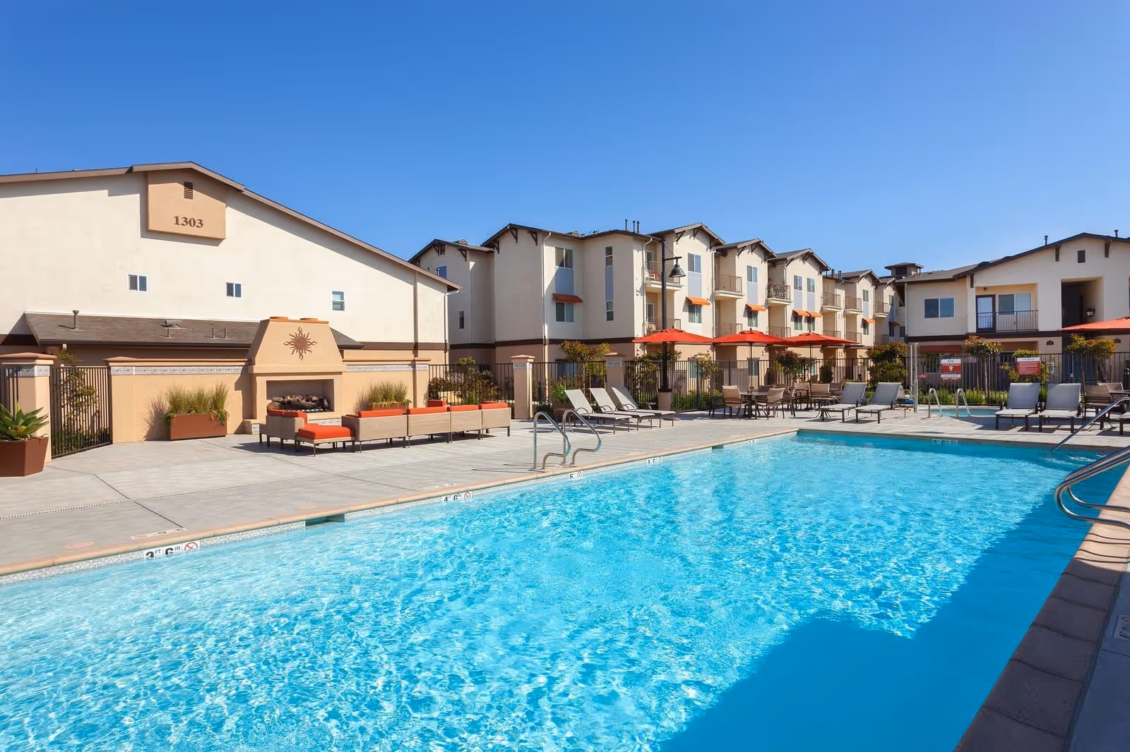 Outdoor swimming pool with lounge chairs, red umbrellas and a multi-story residential building in the background.