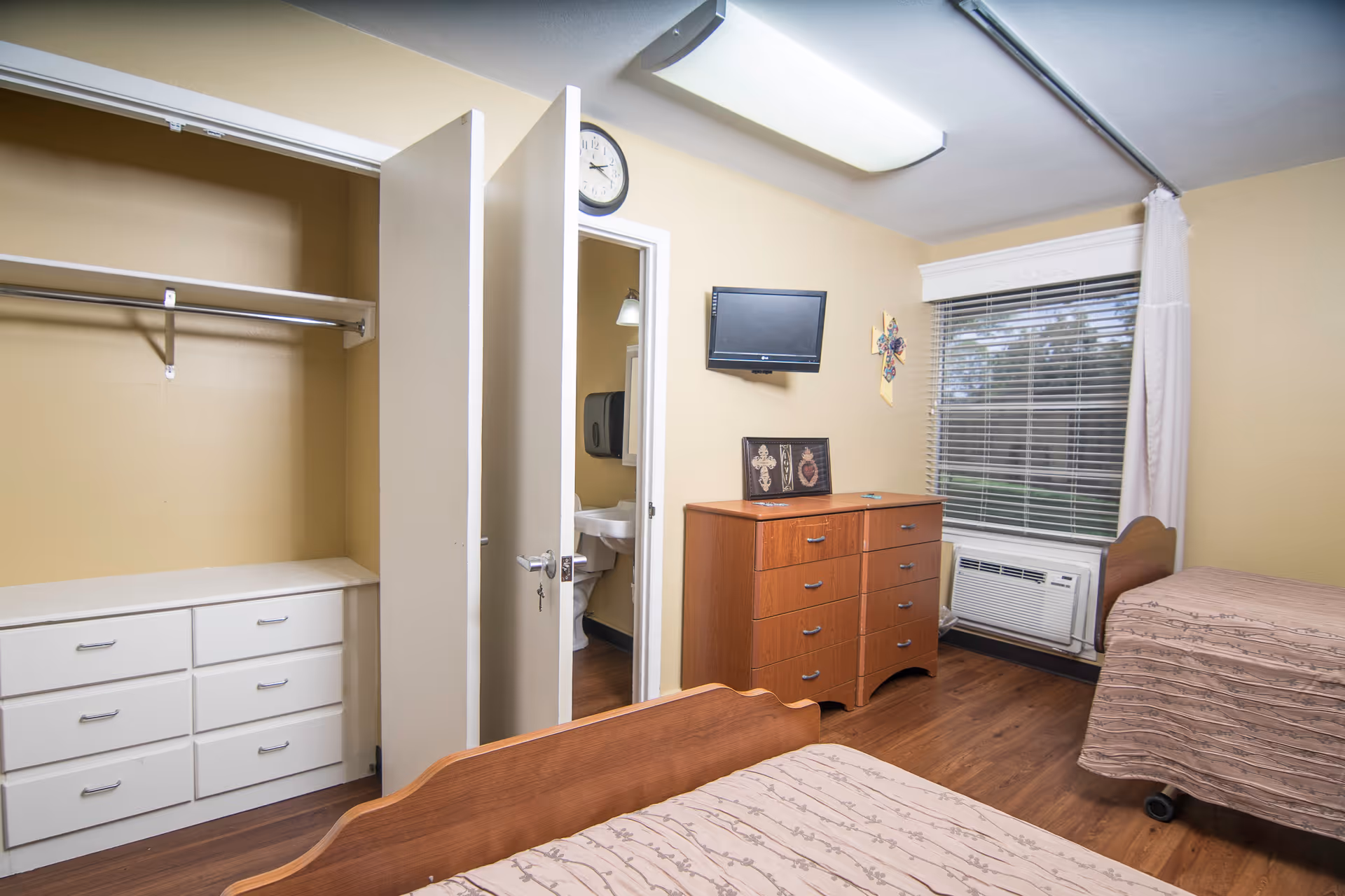 A senior living bedroom with two single beds covered in beige patterned bedspreads, a wooden dresser with a small TV mounted above it, a window with blinds and a white curtain, an open closet with drawers and a hanging rod, and a partially open door revealing a bathroom with a sink and toilet.