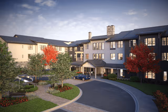 Exterior view of a senior living facility named Balfour Longmont, showing a multi-story building with a covered entrance, surrounded by landscaped greenery including trees with red and green leaves, a curved driveway, and a parked car.