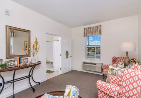 A cozy living room area with two patterned armchairs and a small table with a lamp and flowers. There is a window with a striped valance and an air conditioning unit below it. A console table with framed photos, books, and a vase with yellow flowers is against one wall, and a door leads to a bathroom with a shower curtain visible.
