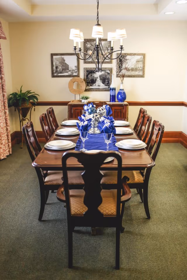 A formal dining room with a long wooden table set for eight people. The table is decorated with a blue runner, blue napkins folded in glasses, white plates, and a centerpiece with silver decorative spheres. There is a chandelier hanging above the table, and a sideboard against the wall with blue and white vases and framed black and white photographs.
