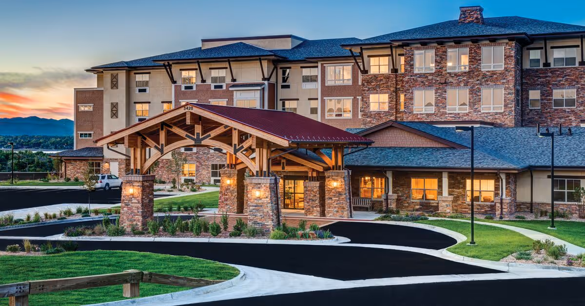 Exterior view of a large senior living community building at dusk with a covered entrance supported by stone pillars and wooden beams, well-maintained landscaping, and a curved driveway.