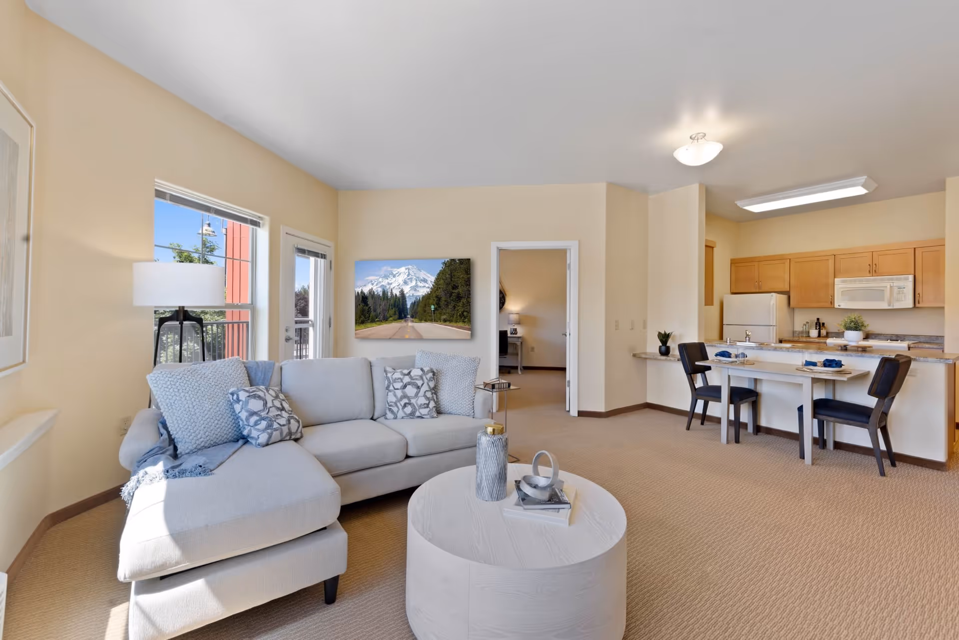 Open-plan senior living apartment interior showing a bright living room with a sectional sofa and coffee table, plus a dining area and kitchen.