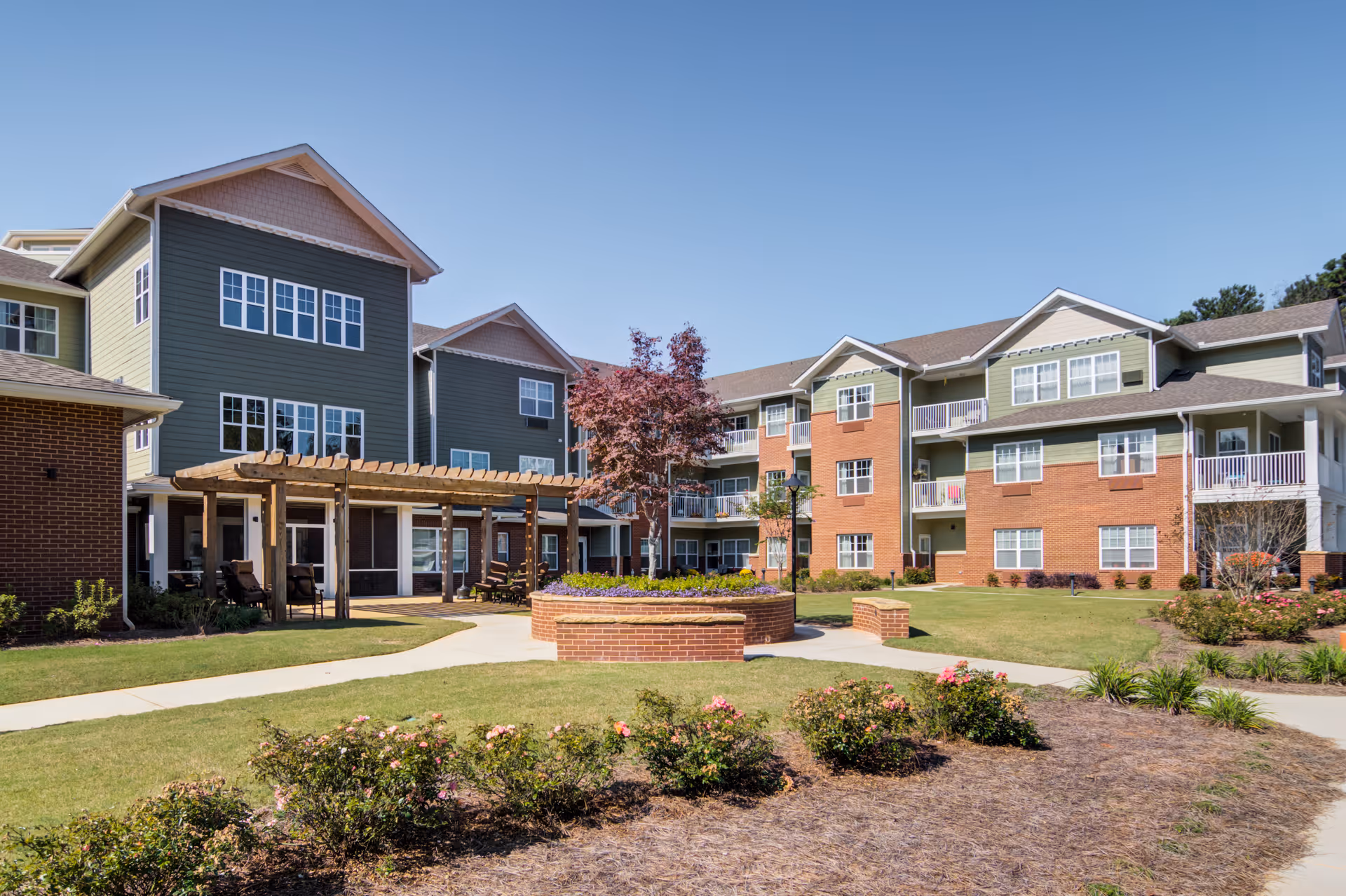Exterior courtyard and front of a multi-story senior living building with landscaped grounds, a pergola, and a circular brick planter.