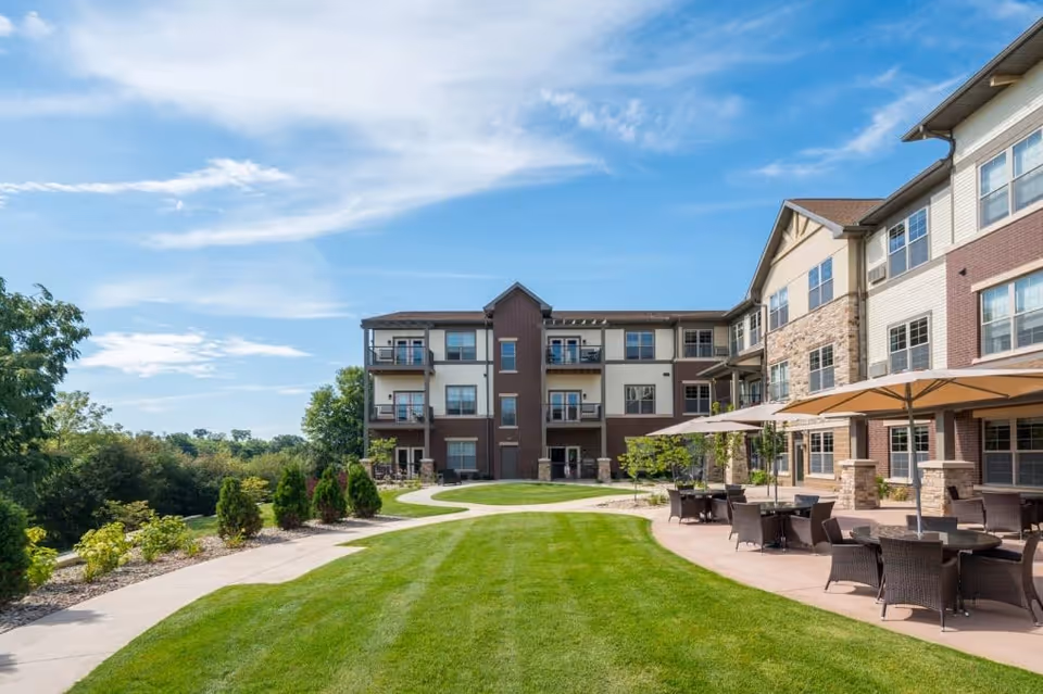 Outdoor view of Noel Manor Retirement Living showing a well-maintained lawn with a paved walkway, patio area with tables, chairs, and umbrellas, and a multi-story building with balconies and large windows under a partly cloudy blue sky.