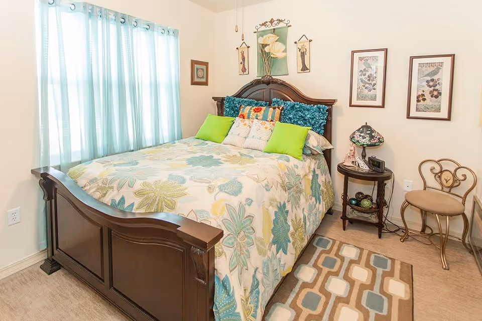 A cozy bedroom with a dark wooden bed frame, a floral patterned bedspread in shades of green, blue, and yellow, and multiple decorative pillows including bright green ones. The room has light-colored walls with framed artwork and a small round side table holding a Tiffany-style lamp, a phone, and other small items. A window with light blue curtains allows natural light to enter, and a patterned rug lies on the floor next to the bed. There is also a decorative metal chair with a cushioned seat.