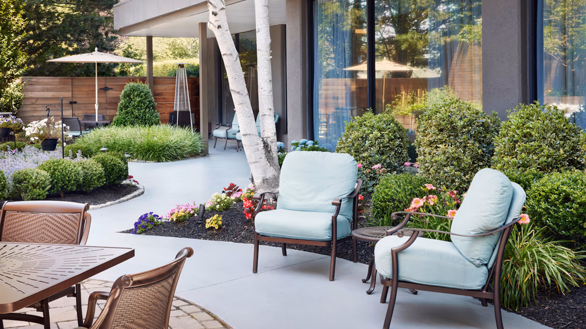 Outdoor patio area with cushioned chairs, a metal table with chairs, surrounded by well-maintained bushes, flowers, and trees next to a building with large windows.