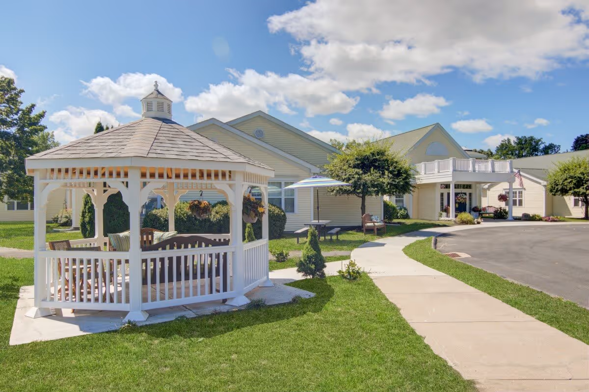 Outdoor view of The Atrium at Cardinal Drive featuring a white gazebo with seating, a paved walkway, green lawn, trees, and a light yellow building under a partly cloudy blue sky.