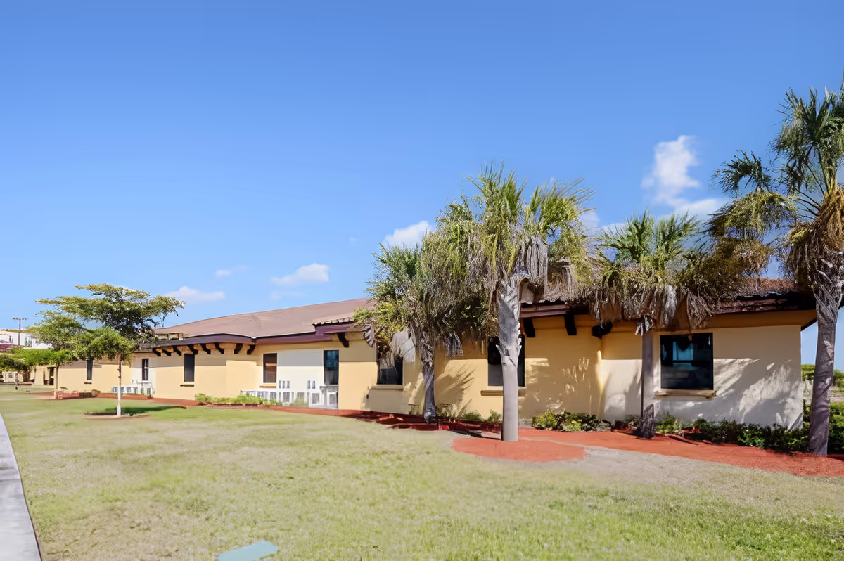 Exterior view of a single-story building with a tan facade and brown roof, surrounded by green grass, palm trees, and a clear blue sky.