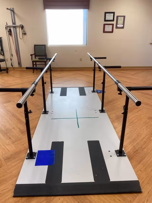 Physical therapy room with parallel bars mounted on a platform for walking exercises, a chair against the wall, exercise equipment on the left, and framed certificates on the wall near a window with a closed blind.