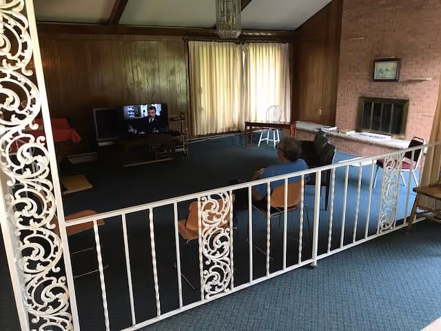 Carpeted communal living room with wood-paneled walls, a TV, fireplace, chairs, a decorative white metal railing, and a person seated facing the television.