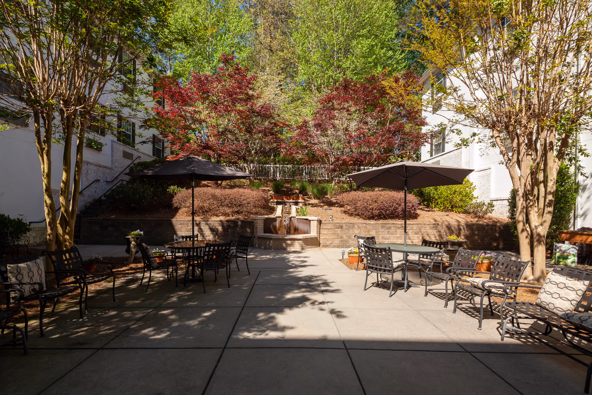 Outdoor patio area with metal tables and chairs, some shaded by large umbrellas. The patio is surrounded by trees with green and red leaves, and there is a small water fountain feature built into a raised brick planter at the back. The area is well-lit with sunlight and has a peaceful garden atmosphere.
